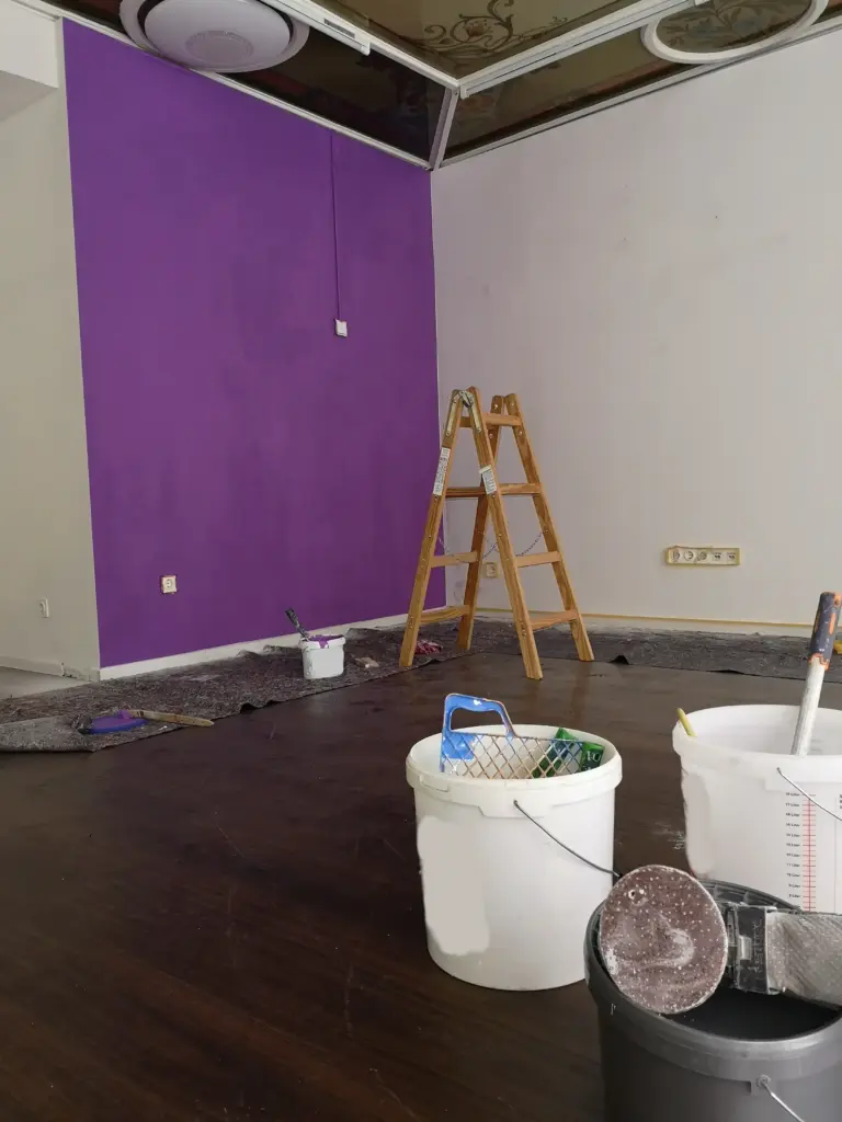 Im Vordergrund sieht man Malerausrüstung im Geschäft, im Hintergrund eine Treppenleiter; die erste Wand ist lila gestrichen.